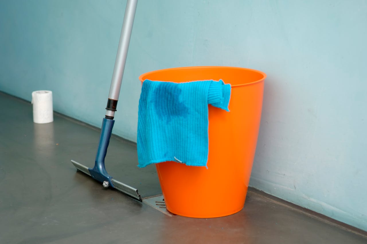 hero-img Close-up of cleaning supplies including orange bucket, mop, and blue rag indoors.